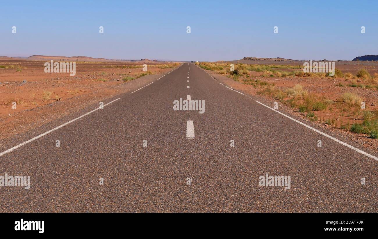 Vue centrale de la route pavée de campagne menant à travers le paysage rural et clairsemé du désert près de Taghroute dans le sud du Maroc, l'Afrique avec le scooter d'enlèvement. Banque D'Images