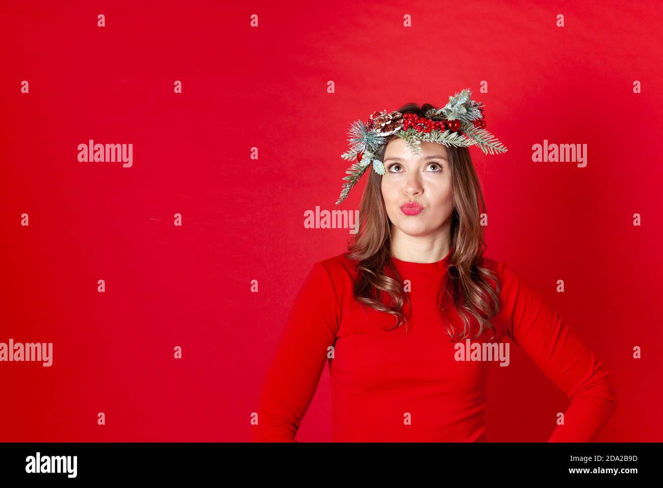 Gros plan d'une jeune femme réfléchie dans une couronne de Noël, les yeux regardant vers le haut, les lèvres comme le canard, sur un fond rouge Banque D'Images