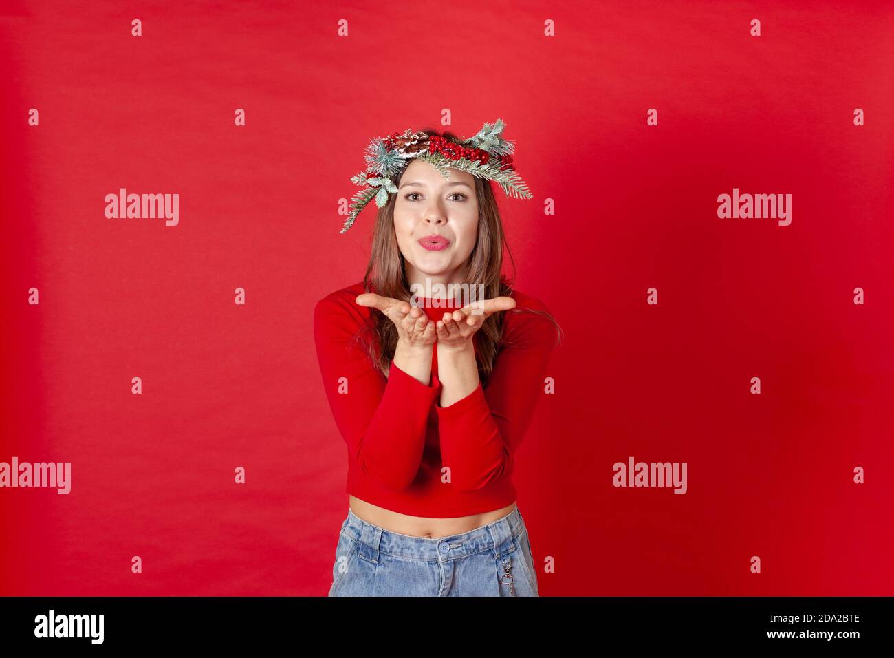 Gros plan d'une jeune femme dans une couronne de Noël, soufflant un baiser sur ses paumes, sur un fond rouge Banque D'Images