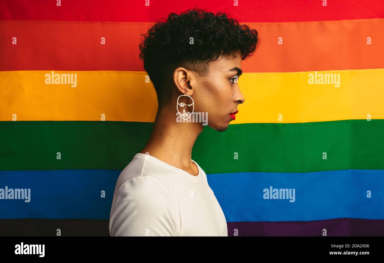 Vue du profil de l'homme gay debout contre le drapeau de fierté. Homme transgenre portant un contour d'oreille et un maquillage. Banque D'Images