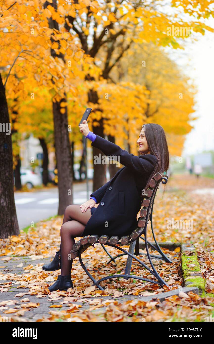 Portrait d'une jeune femme en automne. Décor avec lumière chaude et feuilles en chute. Elle prend un selfie avec le smartpohne. Banque D'Images