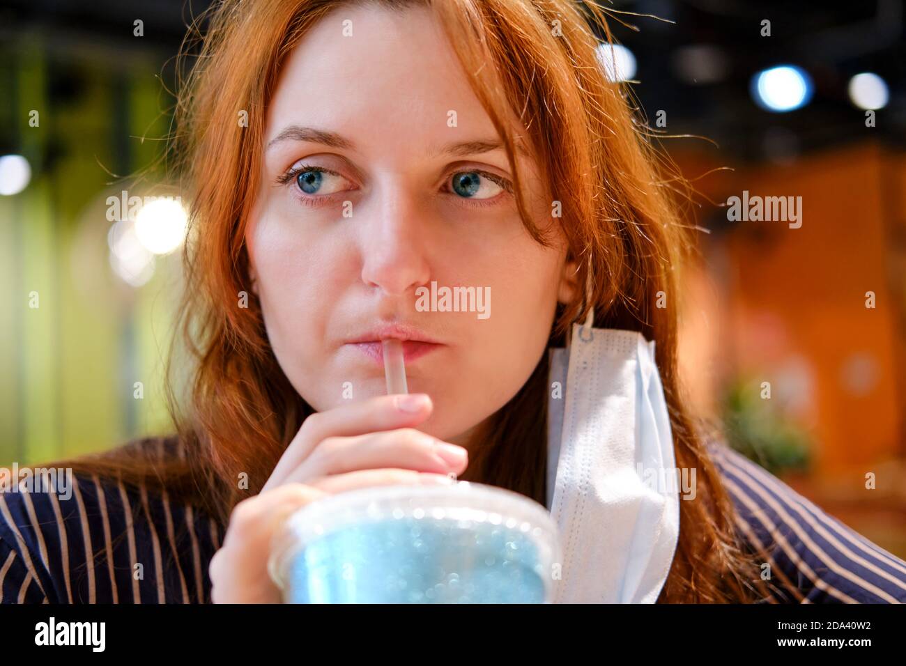 Une jeune femme aux cheveux rouges, dont le masque médical est retiré, est en train de boire un verre dans un café vide. Banque D'Images