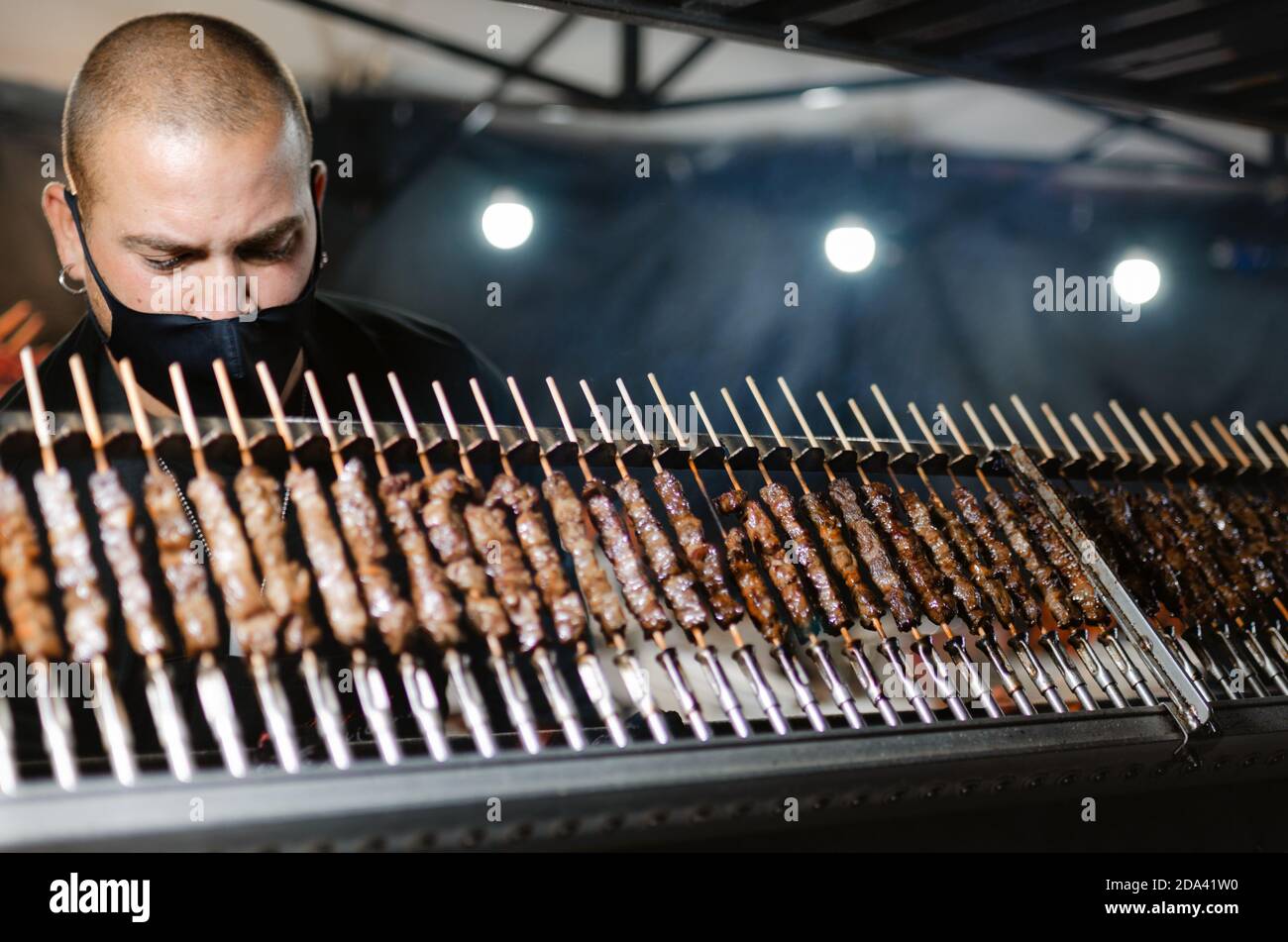 Cuneo, Italie - 3 octobre 2020: Arrosticini traditionnel des abruzzes (brochettes tipycales) cuit la nuit par un jeune homme portant un masque de protection sur une stre Banque D'Images