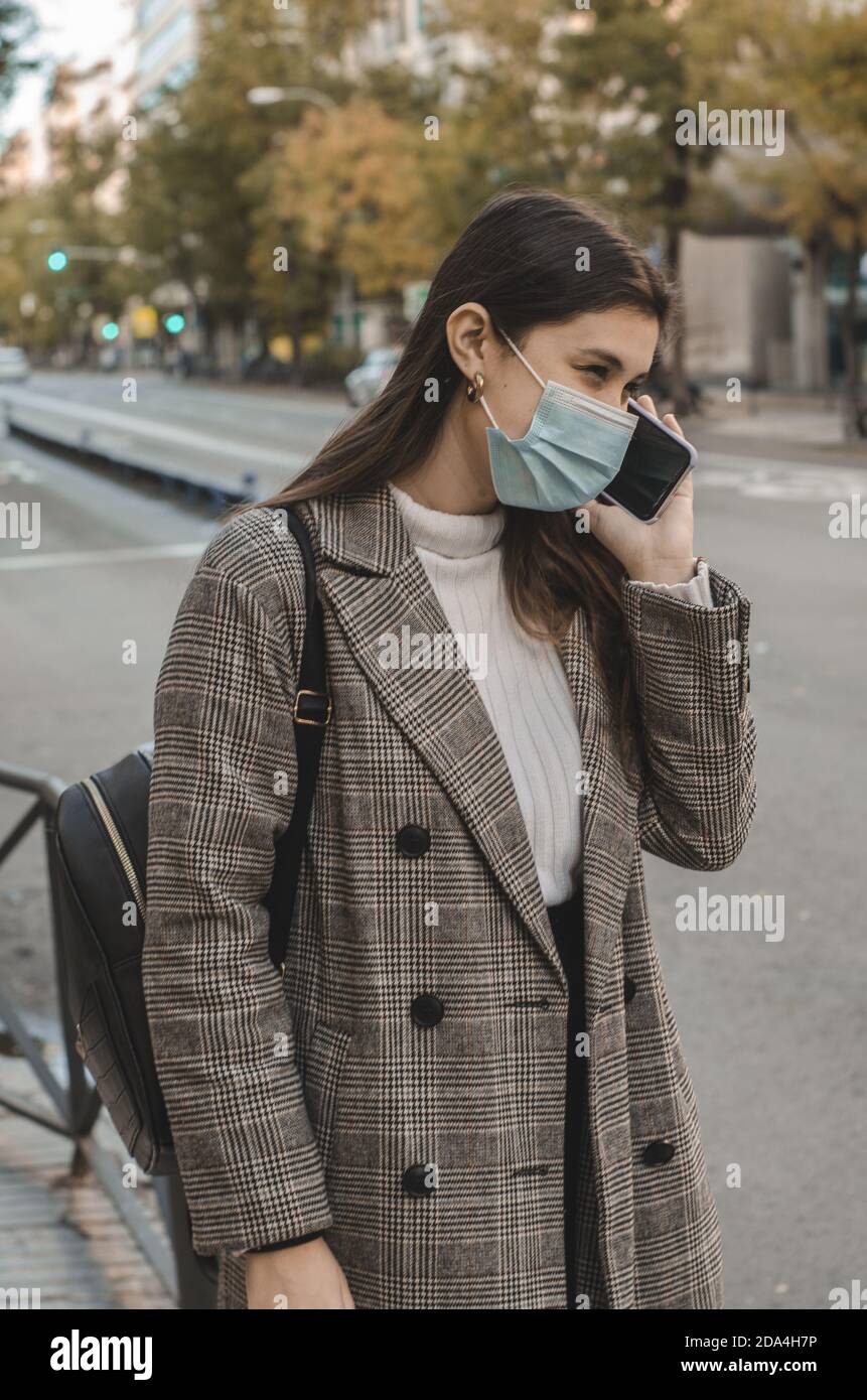 Femme dans un masque facial et une veste à carreaux parlant sur son téléphone tout en marchant dans la rue Banque D'Images