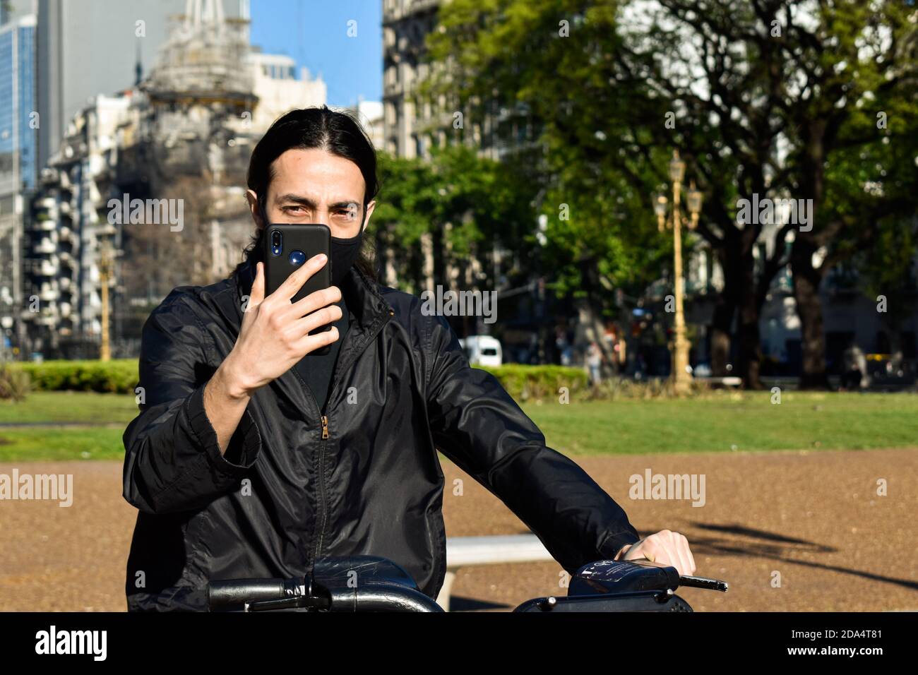 jeune homme à vélo dans un parc municipal regardant son téléphone portable, portant un masque Banque D'Images