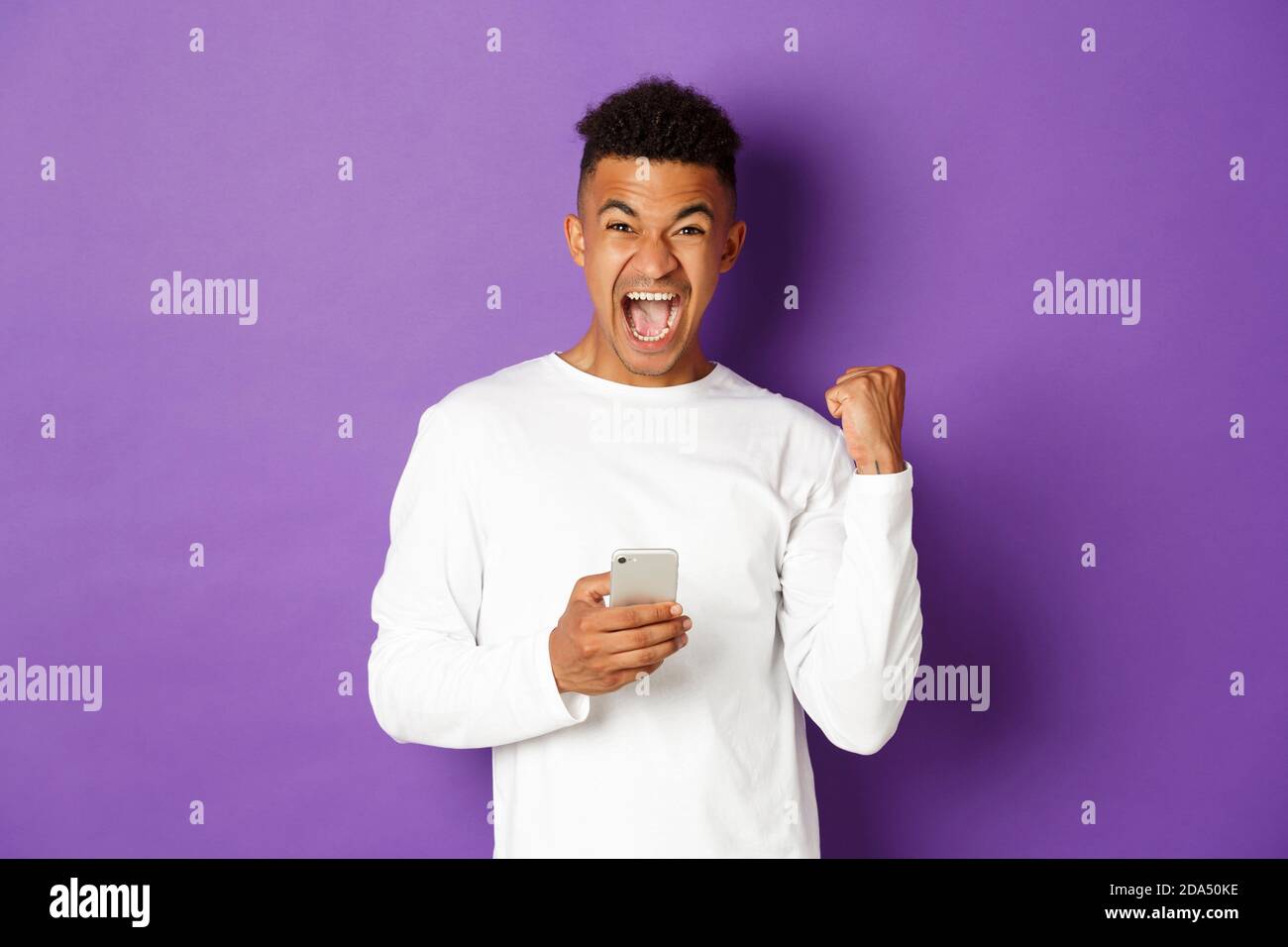 Image d'un homme afro-américain heureux, gagnant quelque chose en ligne, tenant le téléphone mobile et triomphant, en disant oui avec joie, debout sur le violet Banque D'Images
