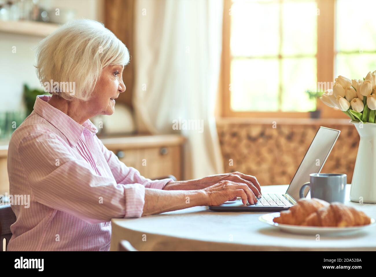 Vue latérale d'une femme âgée travaillant sur un ordinateur portable tout en buvant du thé le matin à la maison. Concept de style de vie domestique. Copier l'espace Banque D'Images