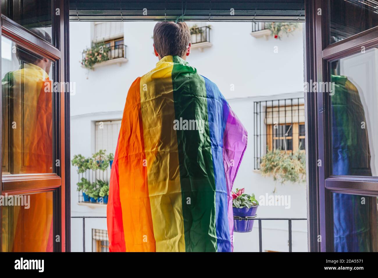 Garçon debout sur un balcon face à la rue avec un drapeau arc-en-ciel sur ses épaules. Lgtbi, gay Banque D'Images