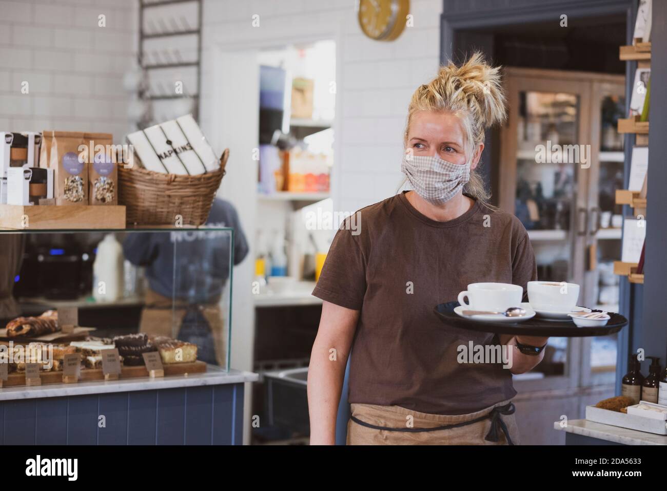 Serveuse blonde portant un masque de travail dans un café, portant un plateau avec des tasses à café. Banque D'Images