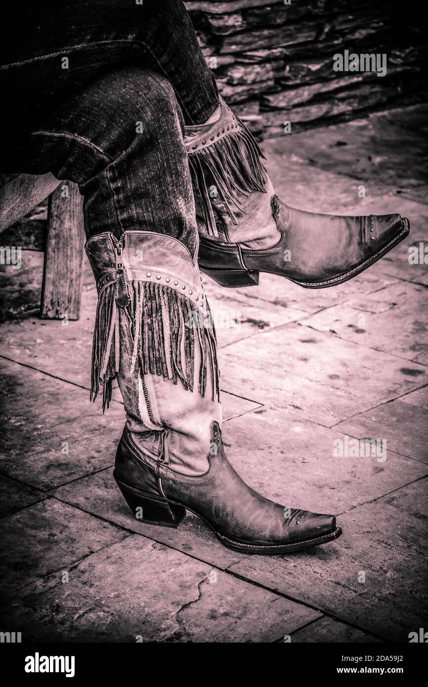 Gros plan sur les jambes de femmes croisées dans des jeans nichés à l'intérieur de bottes de cowgirl à franges sur le trottoir dans le sud des États-Unis, dans des tons chauds noir et blanc Banque D'Images
