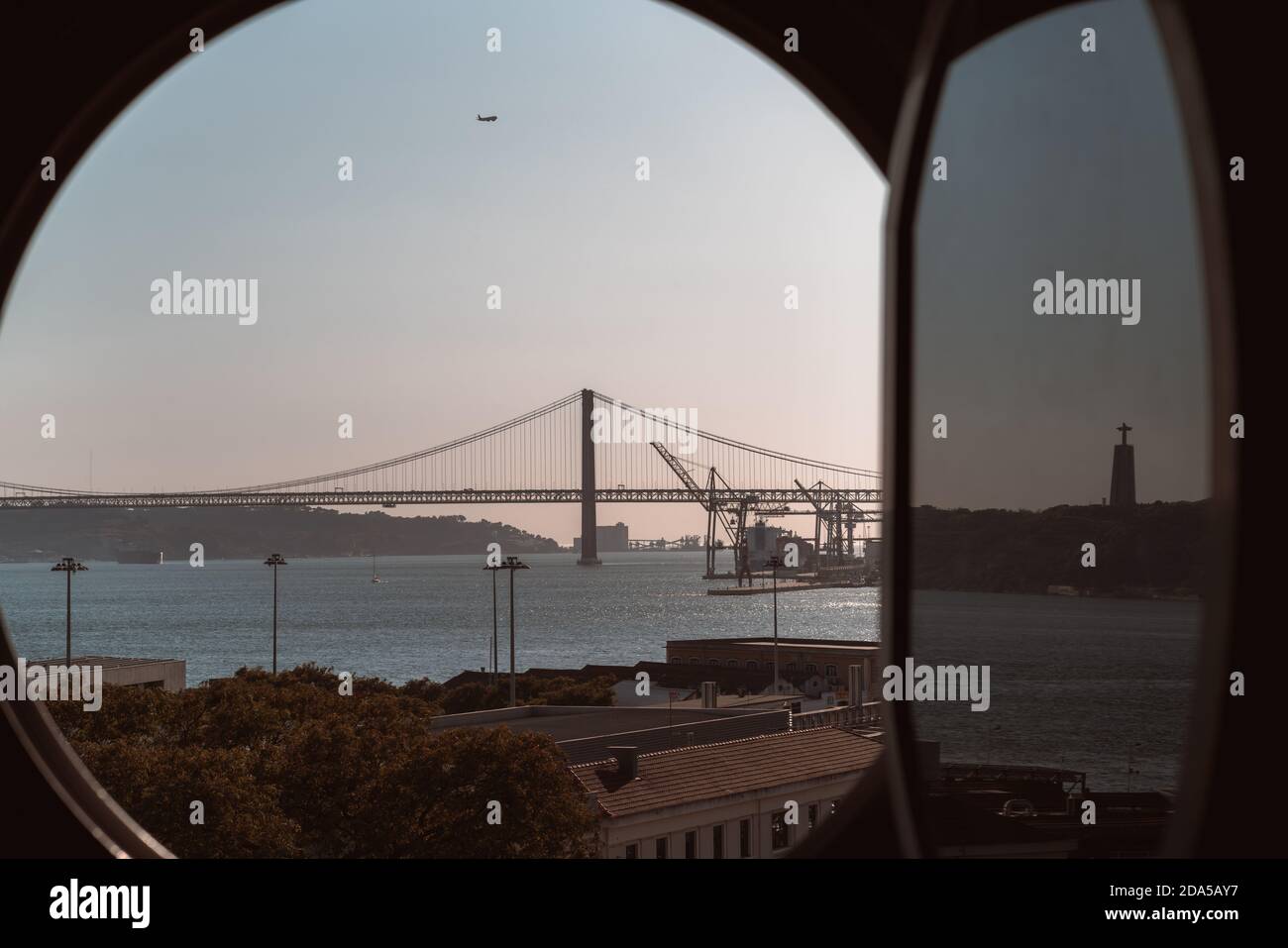 Vue à travers la fenêtre ouverte de l'illuminateur rond d'un ancien historique Construction d'un paysage urbain de Lisbonne en soirée avec un pont La rivière Tejo dans la dista Banque D'Images