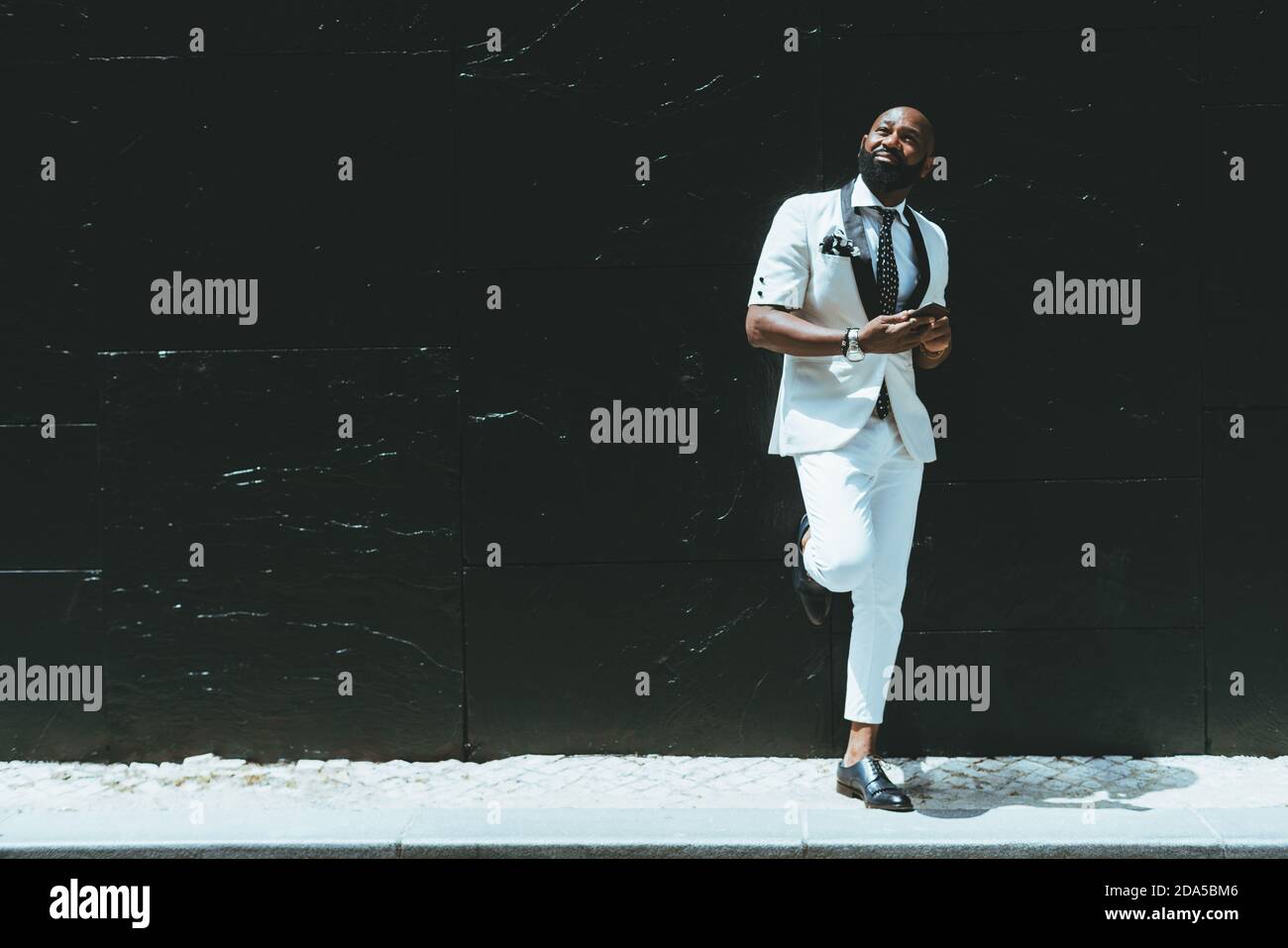 Un élégant et beau chauve barbu afro-américain dans un style à la mode costume blanc avec cravate et smartphone dans son les mains sont penchées contre un Banque D'Images