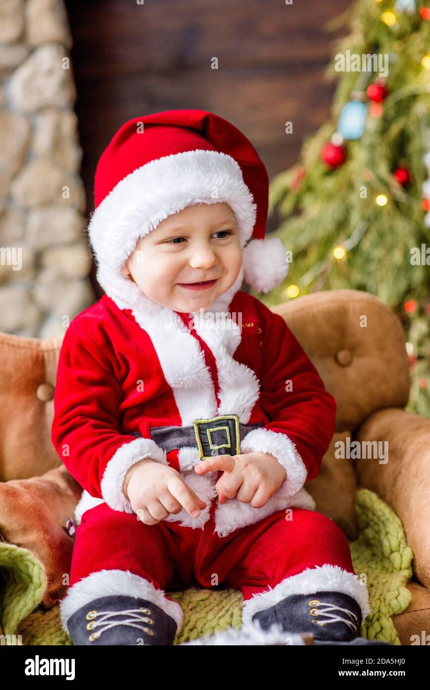 Un petit enfant en costume rouge du Père Noël, assis sur le canapé de la résidence, tenant un cadeau entre ses mains. Carte du nouvel an. Le garçon blond rit. Banque D'Images