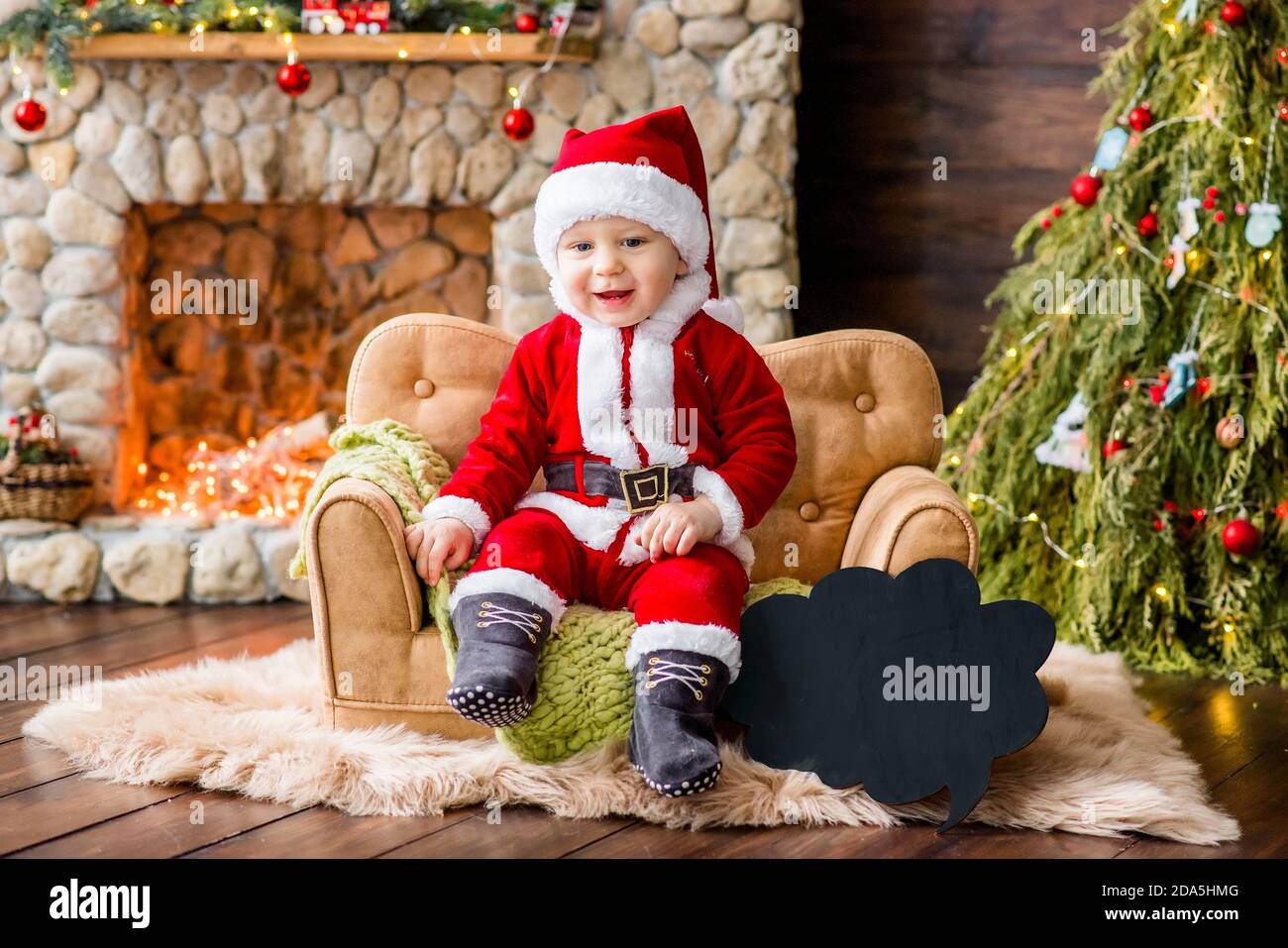 Un petit enfant en costume rouge du Père Noël, assis sur le canapé de la résidence, tenant un cadeau entre ses mains. Carte du nouvel an. Le garçon blond rit. Banque D'Images