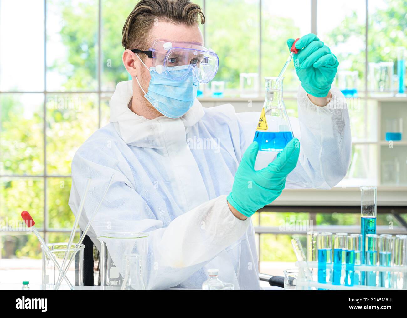 Un scientifique mâle porte une combinaison de protection pour le maintien d'un bécher ou d'un tube avec prélever du liquide à l'intérieur et travailler avec de nombreux équipements de laboratoire pour recherche en laboratoire Banque D'Images