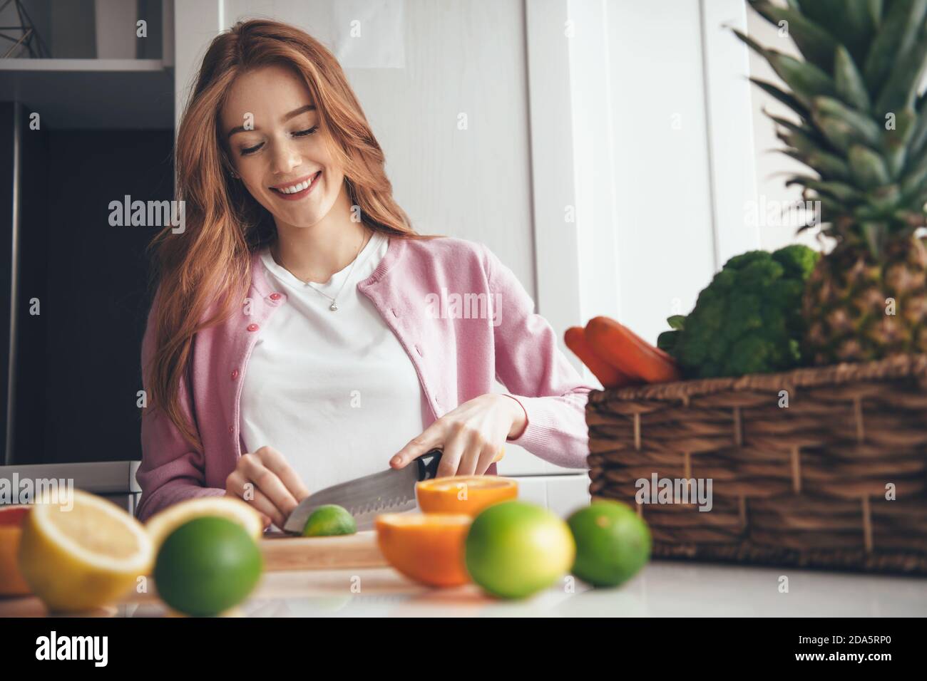 Adorable femme caucasienne avec des taches de rousseur et des cheveux rouges est en tranches un citron vert avec un couteau faisant une salade près d'un panier de fruits Banque D'Images