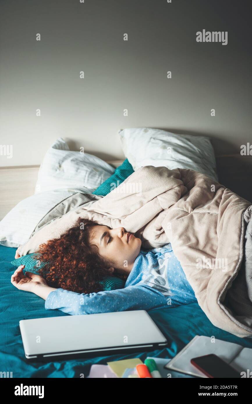 Femme caucasienne fatiguée avec des cheveux bouclés se détendant après avoir fait ses devoirs sur l'ordinateur portable dans le lit Banque D'Images