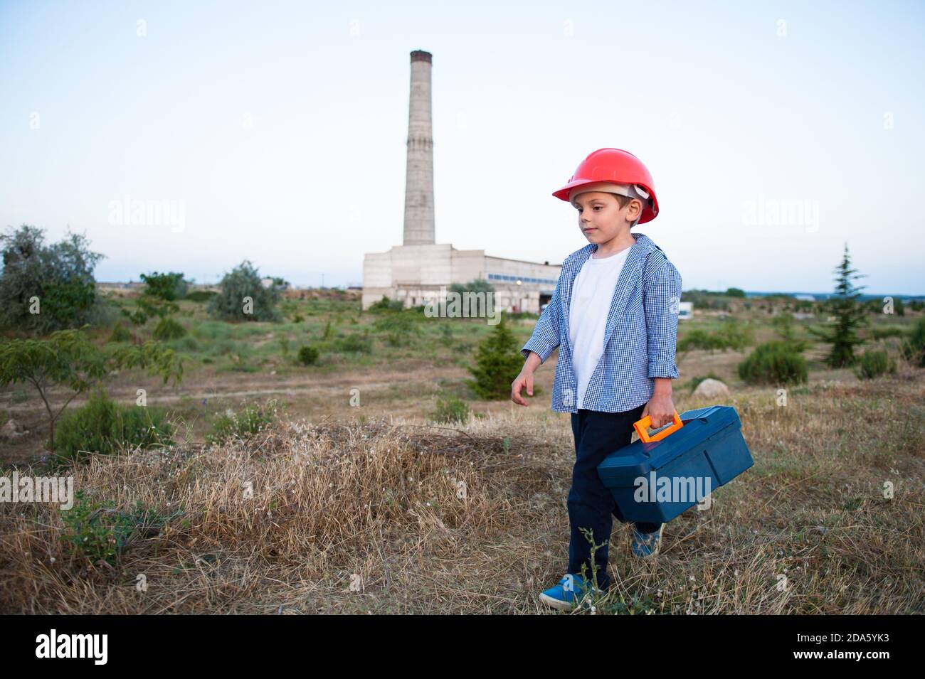 concept de travail d'enfant de petit enfant dans casque orange avec toolbox suit l'usine de l'industrie lourde avec des tuyaux hauts Banque D'Images