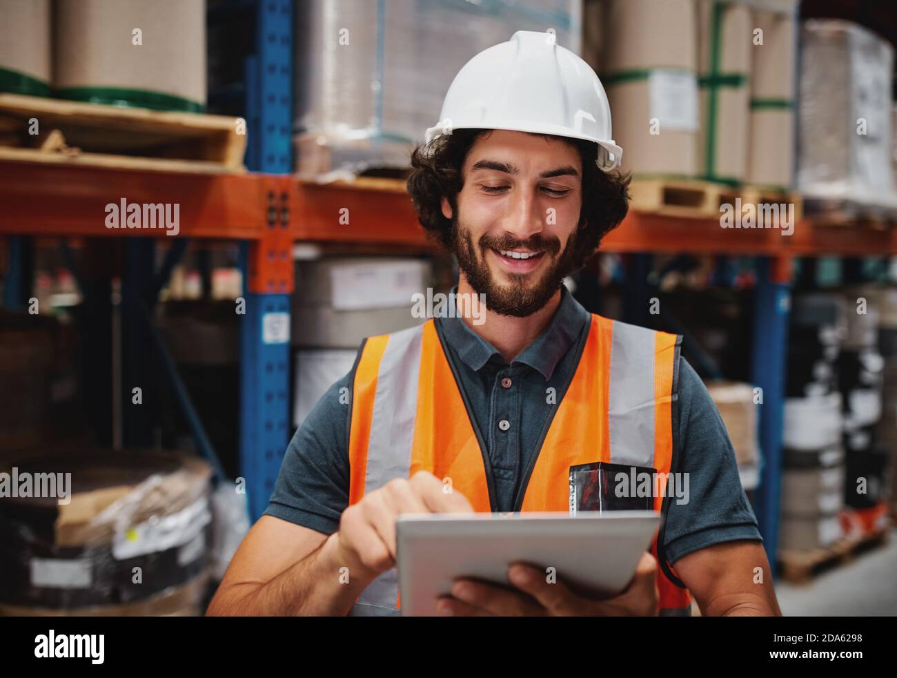 Jeune caucasien souriant travailleur avec casque sur la tête en utilisant tablette pour le travail en se tenant dans l'entrepôt contre des marchandises Banque D'Images