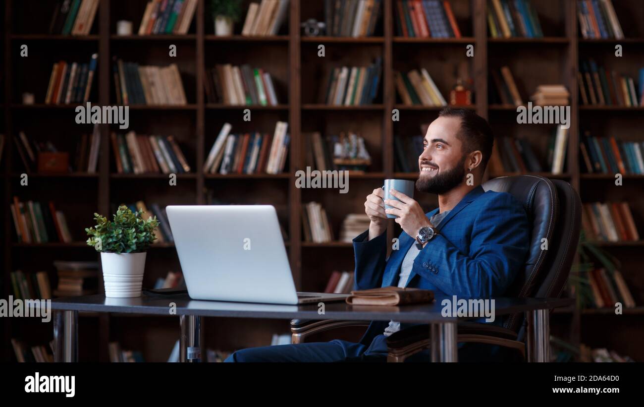 Savourez un café frais au travail. Un jeune homme plein de confiance dans un vêtement élégant et décontracté travaillant sur un ordinateur portable et buvant une boisson chaude tout en étant assis au travail Banque D'Images