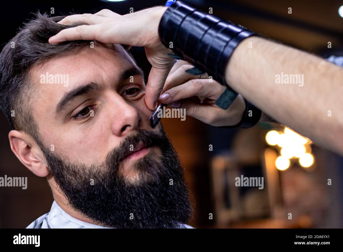 Jeune barbu qui obtient une coupe de cheveux par un professionnel à la  barbershop moderne. Rasage et mise en forme de la barbe et des pattes avec  un rasoir droit vintage. Movember