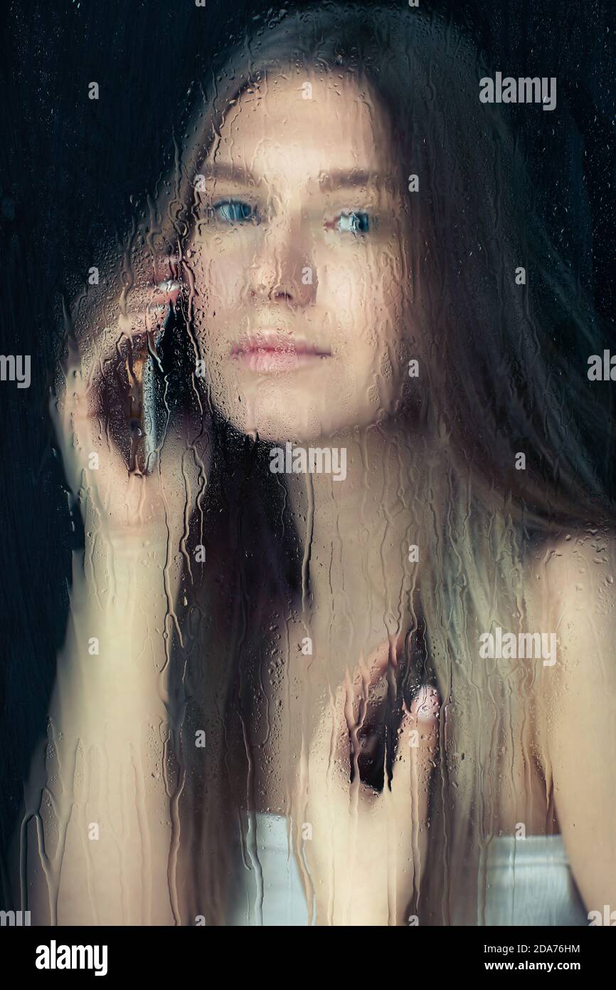 Portrait d'une jeune femme blonde avec les mains du téléphone. Femelle derrière la vitre avec gouttes de pluie. Concept de verrouillage d'auto-isolation. Banque D'Images
