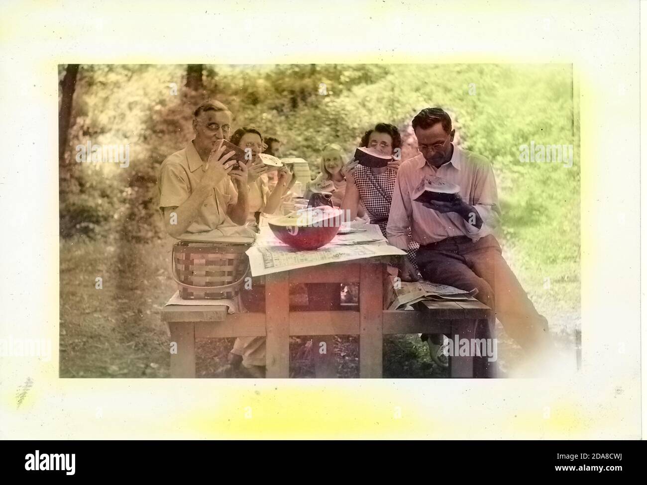 Photographie en noir et blanc, montrant six personnes, y compris des hommes, des femmes et des enfants, assis autour d'une table de pique-nique couverte de journaux, mangeant de gros morceaux de pastèque, avec un panier de pique-nique en osier vintage sur un banc, Et une demi-pastèque sur la table, probablement photographiée en Ohio dans la décennie qui a suivi la Seconde Guerre mondiale, 1950. Remarque : l'image a été colorisée numériquement à l'aide d'un processus moderne. Les couleurs peuvent ne pas être exactes. () Banque D'Images