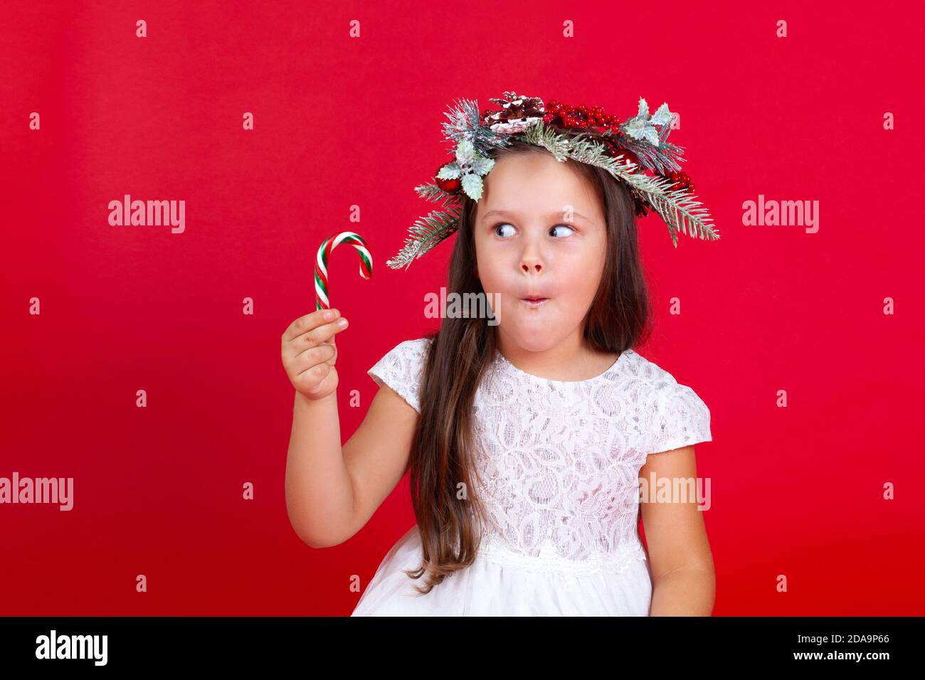 Maquette d'une belle fille dans une couronne de Noël regardant avec surprise la canne au caramel dans sa main, sur un fond rouge Banque D'Images