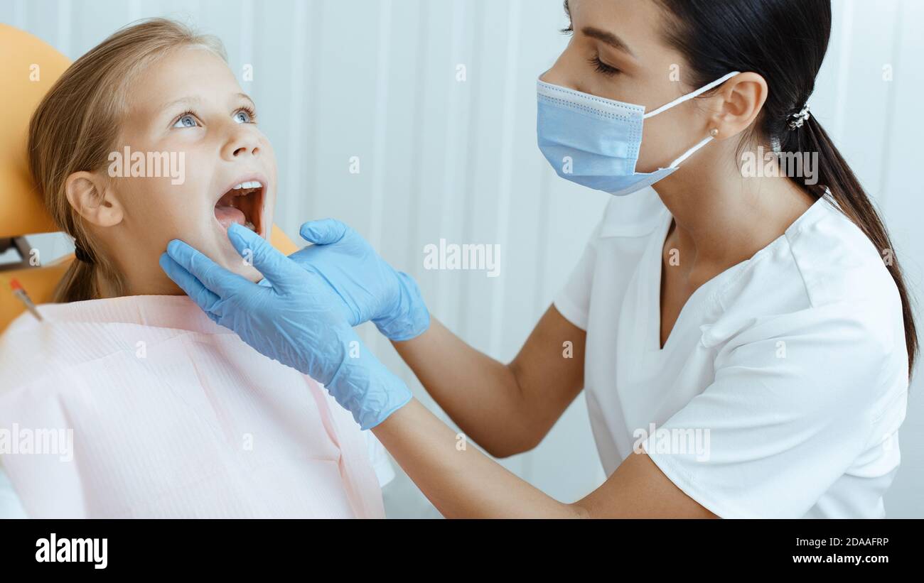 Une jeune femme médecin en manteau blanc, masque de protection et gants examine les dents des enfants Banque D'Images