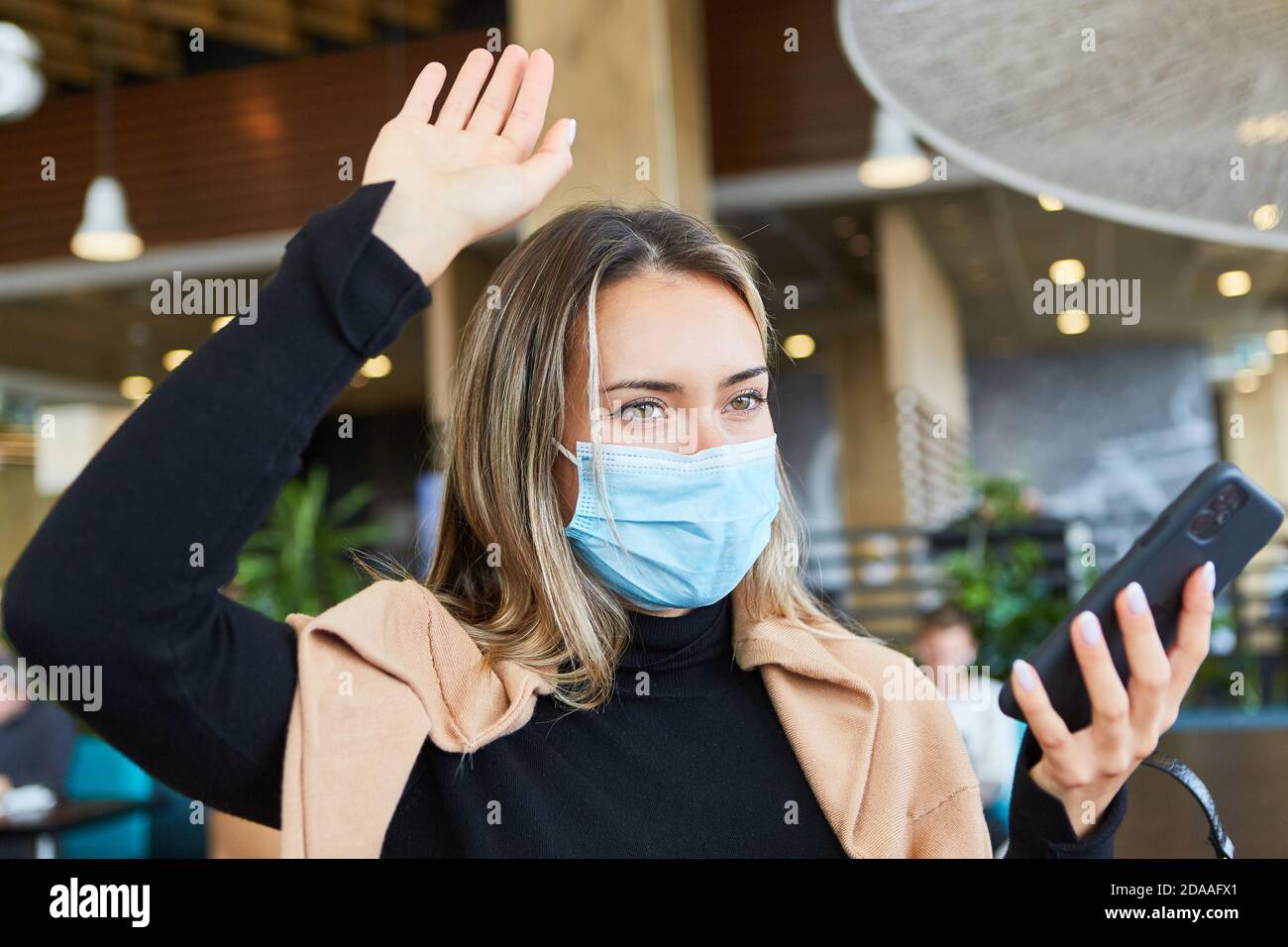 Femme avec masque et smartphone dans un café vagues un accueil à distance Banque D'Images