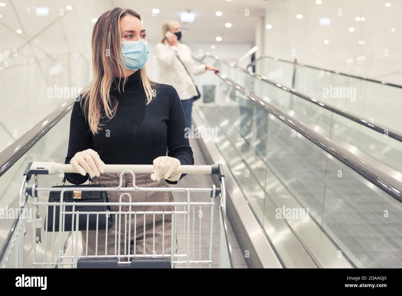 Femme avec masque à cause de Covid-19 avec chariot de shopping sur un escalier roulant de supermarché Banque D'Images