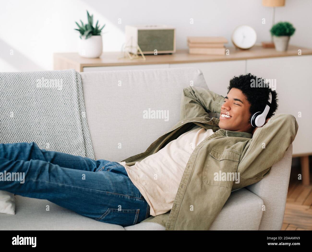 Joyeux adolescent noir dans un casque allongé sur un canapé confortable et à écouter de la musique à la maison, espace de copie Banque D'Images