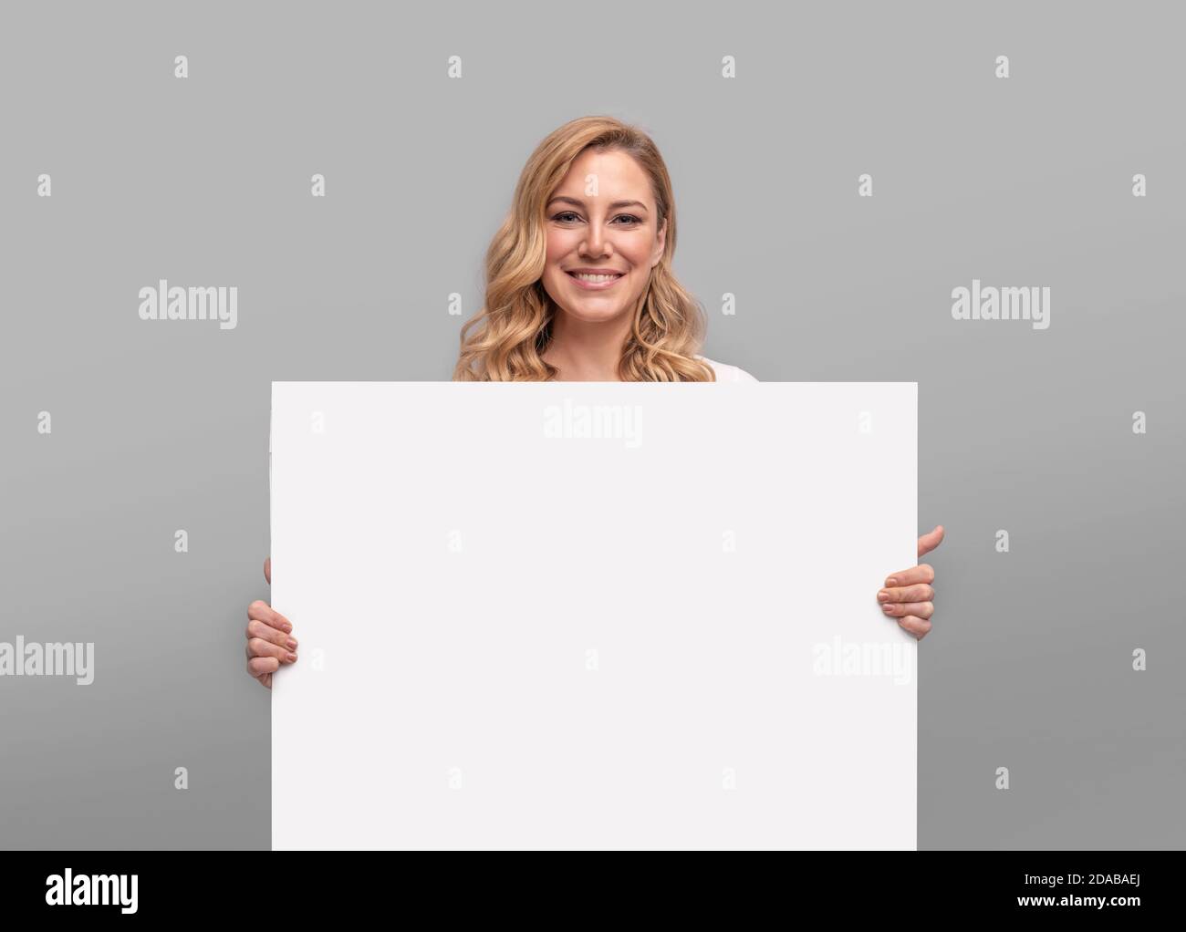 Jeune femme tenant une grande feuille blanche vierge sur un arrière-plan gris Banque D'Images