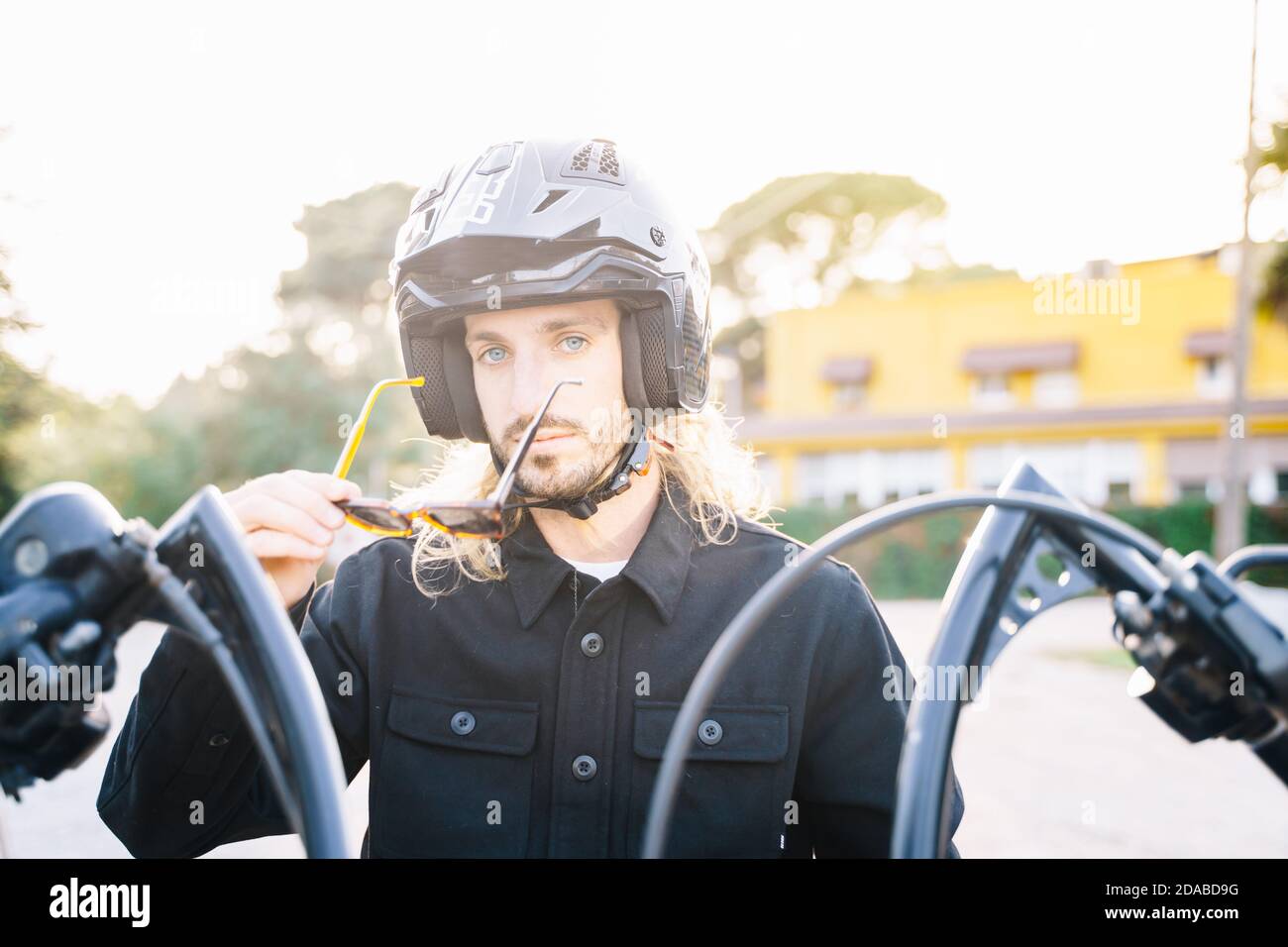 Homme dans un casque qui prend les lunettes de soleil en étant assis dans une moto avec les rayons du soleil derrière lui avec un bâtiment en arrière-plan Banque D'Images