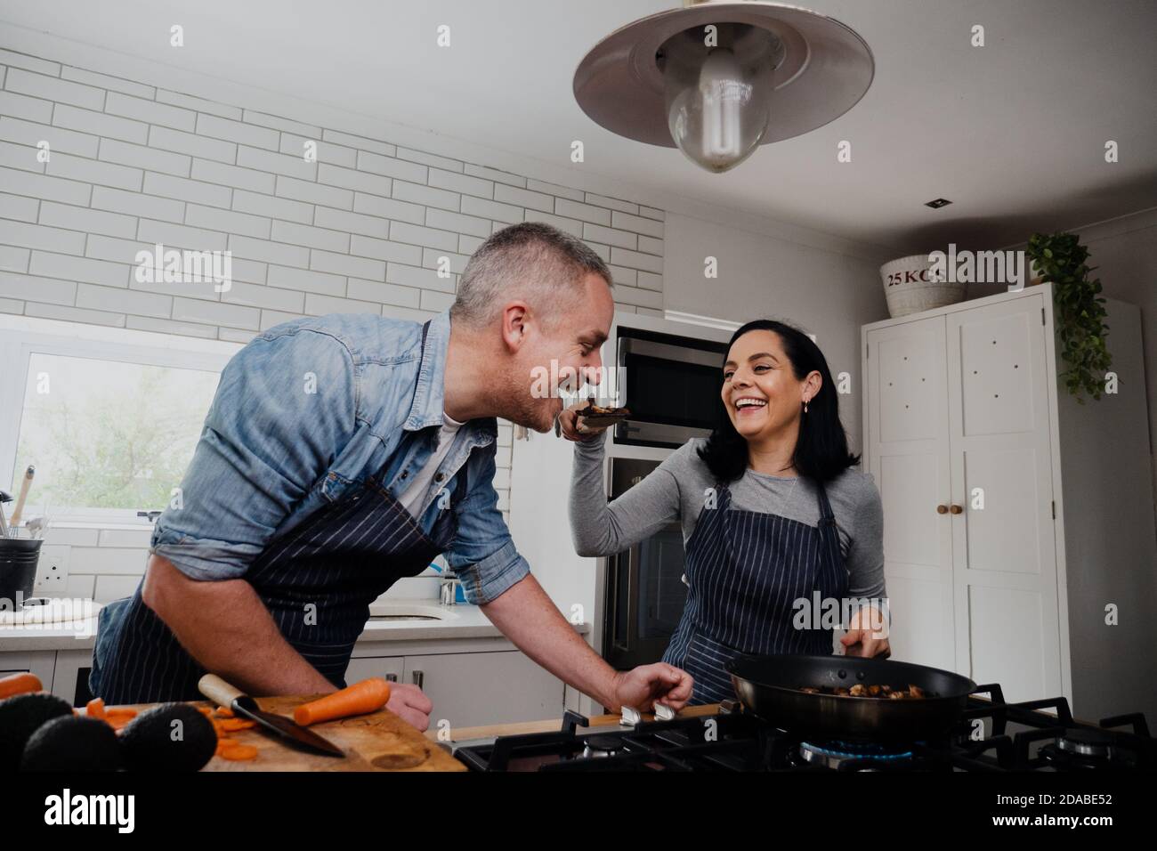 La femme fait goûter à la cuillerée du dîner à son mari tout en préparant le dîner dans une cuisine de luxe. Banque D'Images