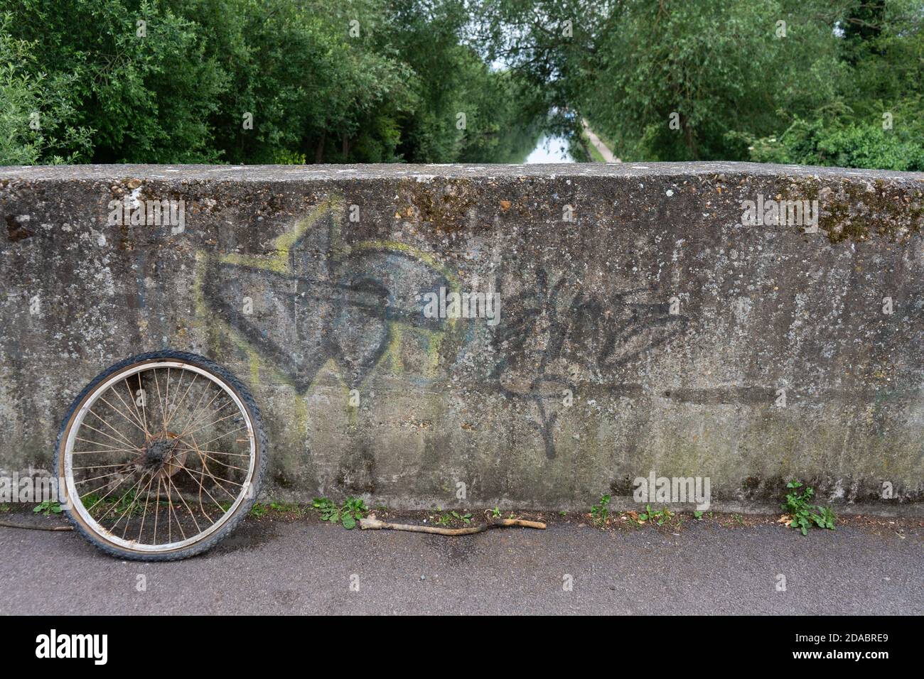 Ancienne roue de vélo rouillée abandonnée sur le pont de graffiti en béton de l'autre côté canal rivière bordée de beaux arbres Banque D'Images