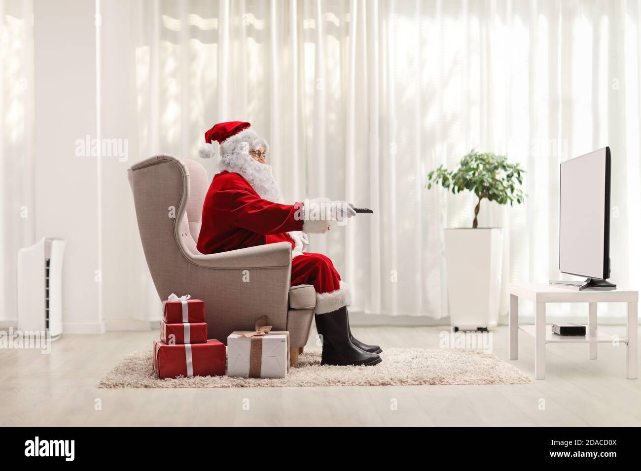 Photo pleine longueur du père noël dans un fauteuil avec une télécommande située devant le téléviseur à la maison avec cadeaux sur le sol Banque D'Images