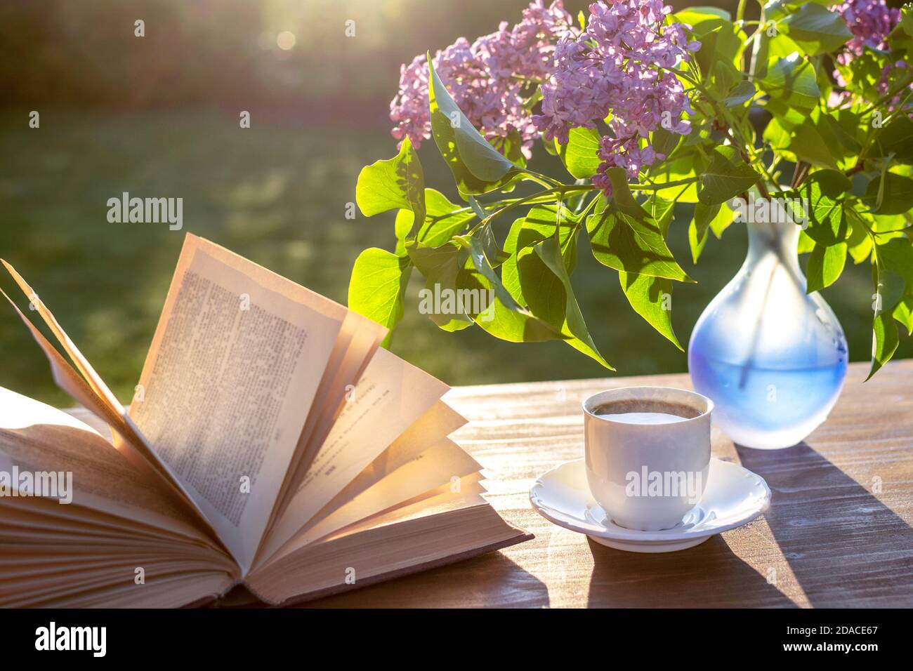 Petite tasse blanche d'espresso, livre ouvert, vase bleu semi-transparent avec fleurs de lilas pourpres sur table rustique en bois dans le jardin Banque D'Images