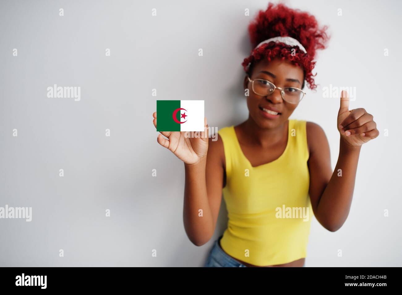 Femme africaine aux cheveux afro, porter un maillot jaune et des lunettes, tenir le drapeau algérien isolé sur fond blanc, montrer le pouce vers le haut. Banque D'Images