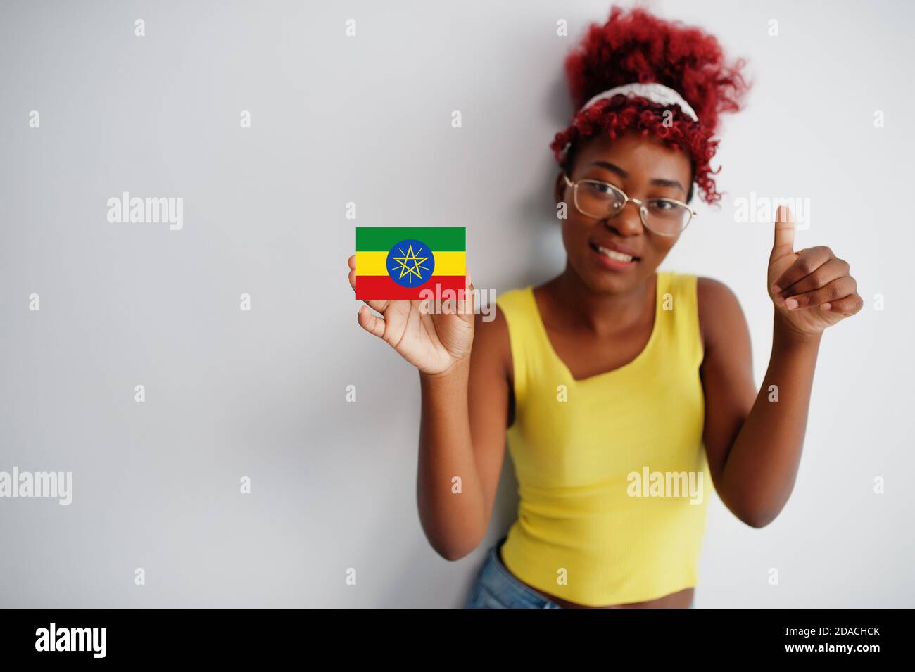 Femme africaine aux cheveux afro, porter un maillot jaune et des lunettes, tenir le drapeau éthiopien isolé sur fond blanc, montrer le pouce vers le haut. Banque D'Images