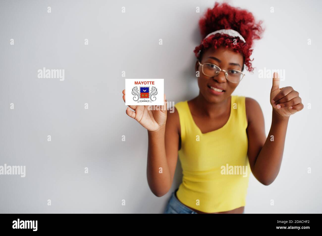 Femme africaine aux cheveux afro, porter un maillot jaune et des lunettes, tenir le drapeau de Mayotte isolé sur fond blanc, montrer le pouce vers le haut. Banque D'Images