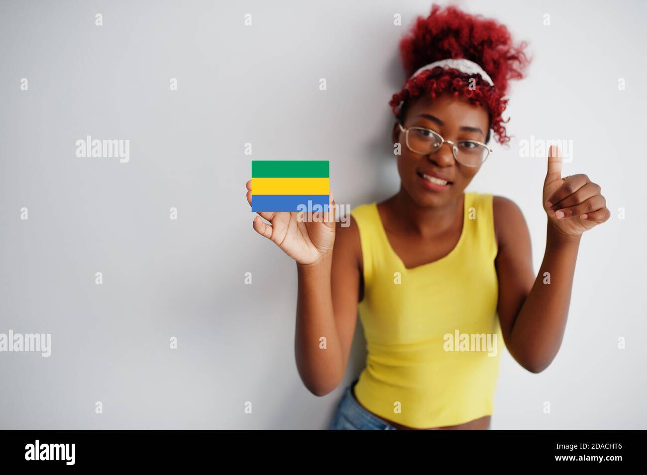 Femme africaine aux cheveux afro, porter un maillot jaune et des lunettes, tenir le drapeau gabonais isolé sur fond blanc, montrer le pouce vers le haut. Banque D'Images