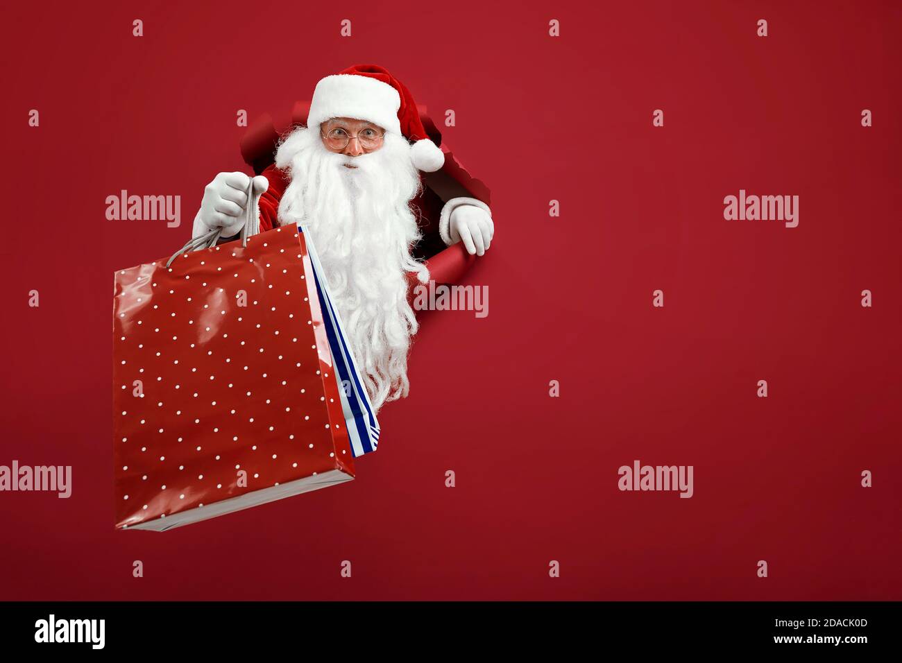 Le père Noël tient le paquet de magasins dans la main par un trou de papier.  Homme barbu dans chapeau de père noël regardant à travers trou sur papier  rouge Photo Stock -