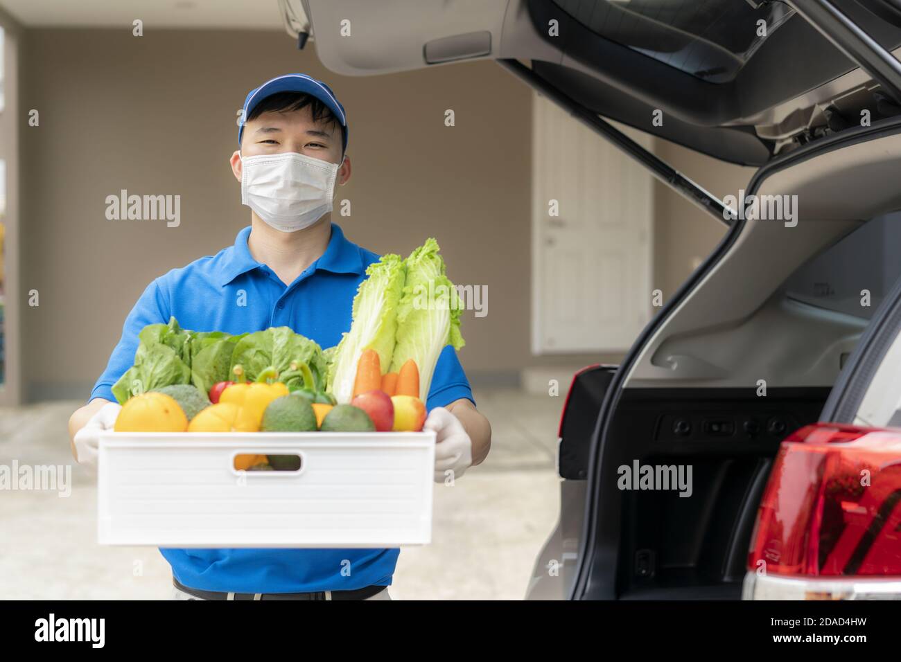 Homme de livraison asiatique portant un masque facial et un gant avec une boîte d'épicerie de nourriture, de fruits, de légumes et de boissons debout près d'une fourgonnette devant la maison de clients Banque D'Images