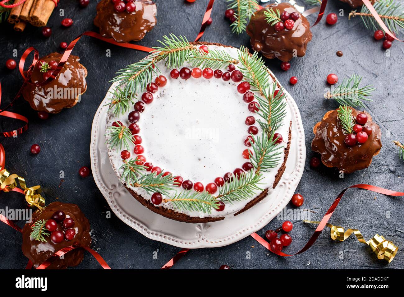 Les petits muffins au chocolat et le grand gâteau sont versés avec du chocolat, décoré de baies d'hiver. Bâtons de cannelle, décoration en flocons de neige sur une table sombre. Plat Banque D'Images