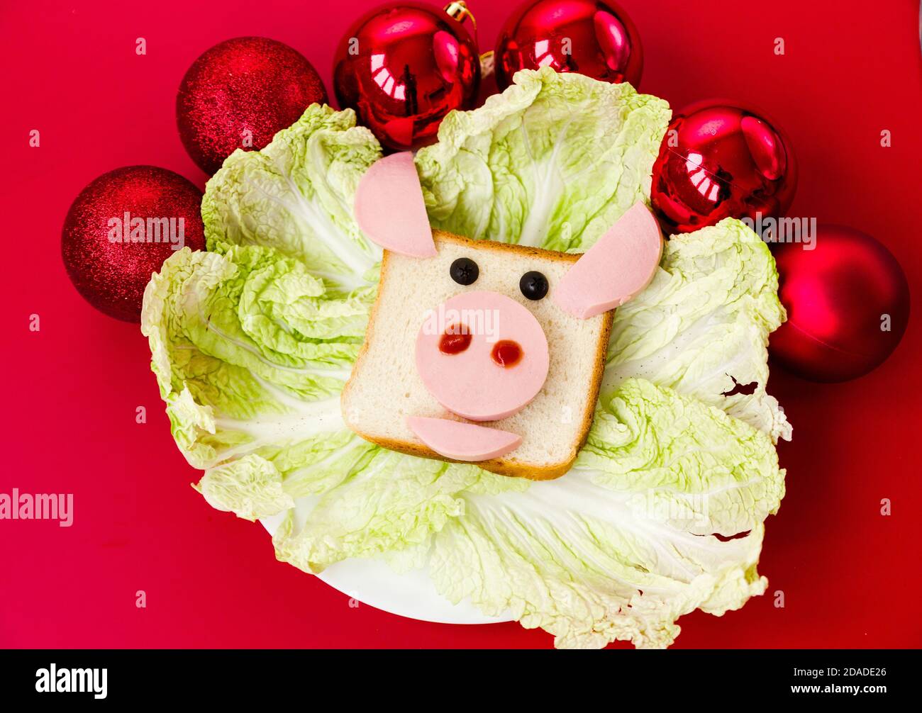 Un sandwich en forme de tête de taureau sur une assiette décorée de moulages de salade. La plaque se trouve sur un arrière-plan rouge. Des boules de noël rouges décorent le Banque D'Images