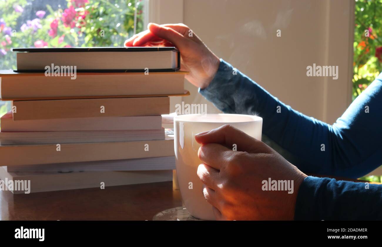 Femme sélectionnant à partir d'une pile de livres assis à un table à la maison avec une tasse de vapeur chaude boisson Banque D'Images