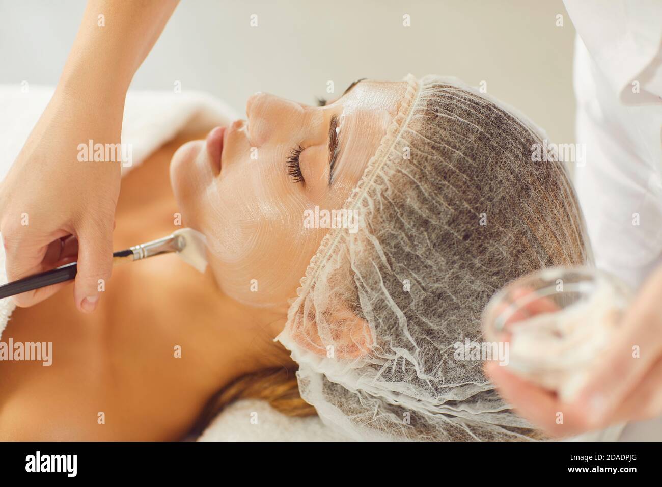 Mains d'esthéticienne appliquant le masque hydratant avec la brosse cosmétique à jeune femme décontractée Banque D'Images