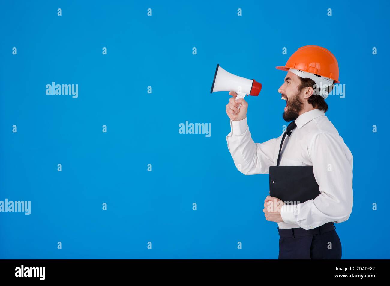 Un jeune homme barbu de construction dans une chemise blanche et un casque de construction sur fond bleu. Un ingénieur-architecte industriel crie Banque D'Images