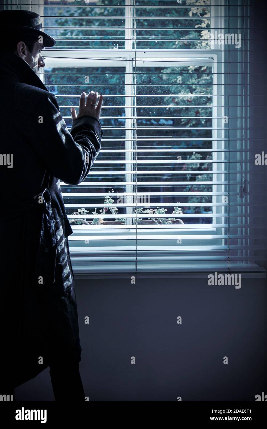 Homme dans le manteau et chapeau Peering dehors stores de fenêtre Banque D'Images